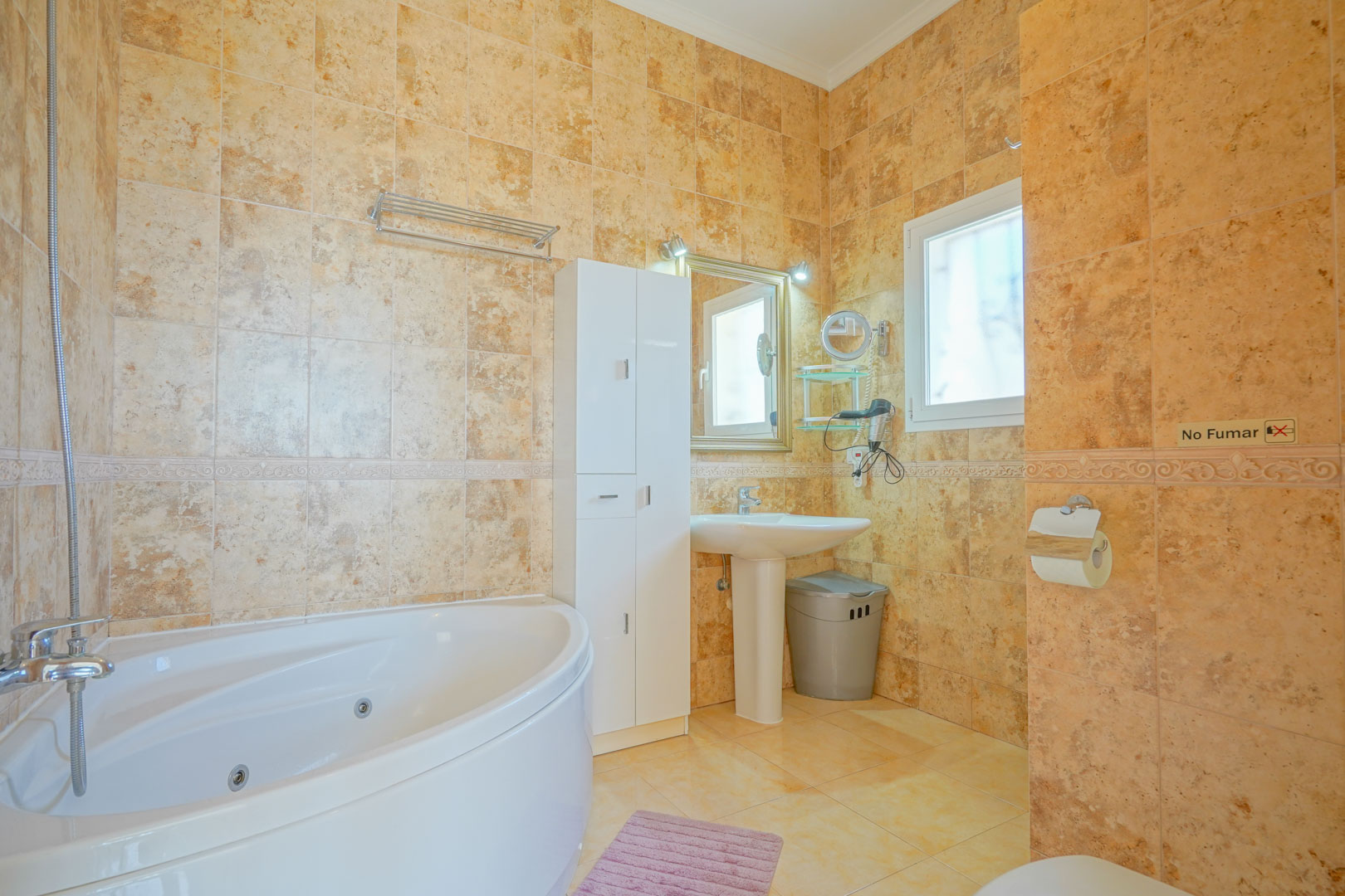 Bathroom featuring a corner whirlpool bathtub, warm-toned ceramic wall tiles, a pedestal sink, and a tall white storage cabinet.