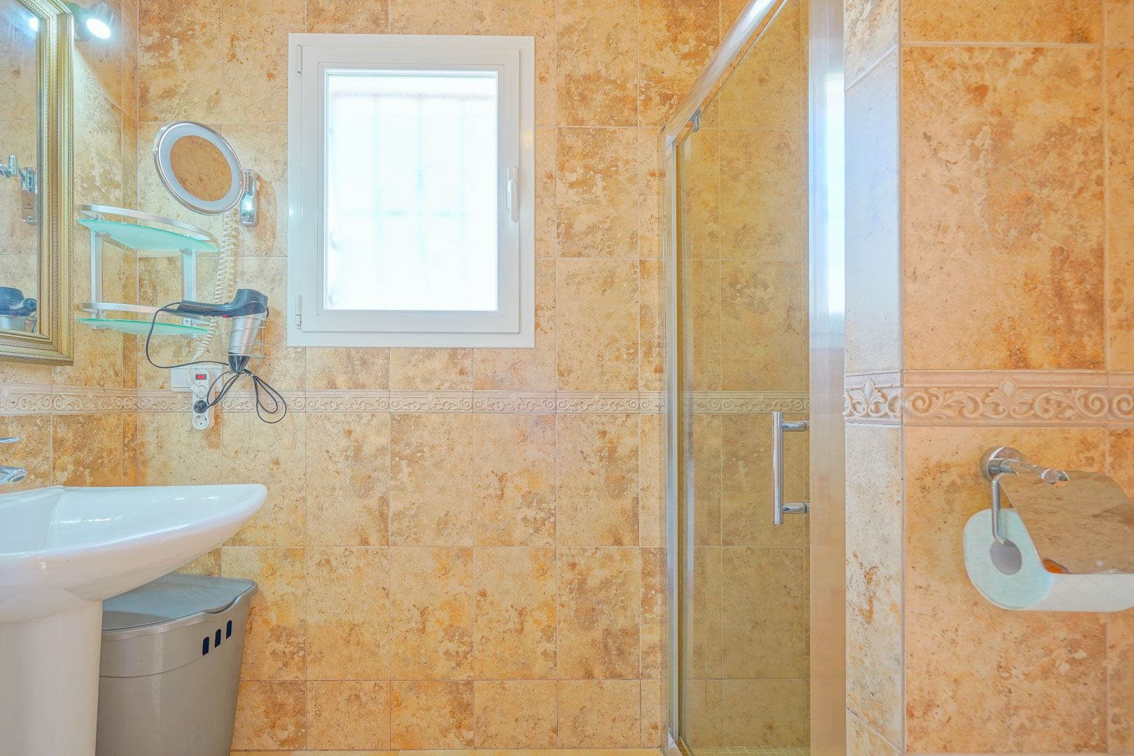 Bathroom featuring earth-toned ceramic tiles, a white pedestal sink, and a glass-enclosed shower. Natural light enters through a frosted tilt-and-turn window.
