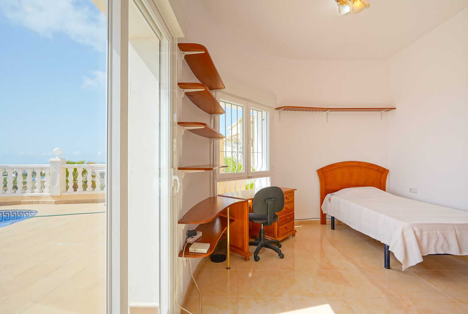 Bedroom featuring a single bed, wooden desk, and integrated shelving. Large sliding glass doors provide direct access to the pool terrace and outdoor area.