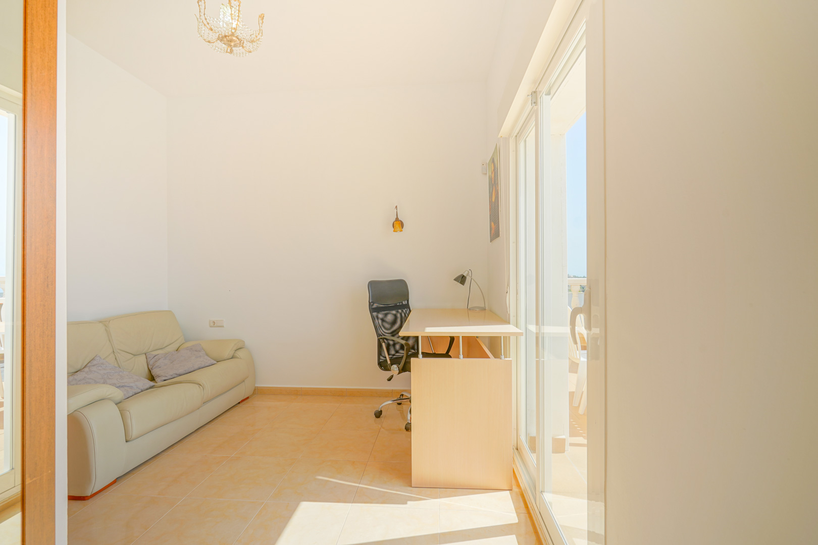 Bright multi-purpose room featuring a desk area, leather sofa, and direct terrace access via sliding glass doors. Natural light highlights the warm ceramic tile flooring.