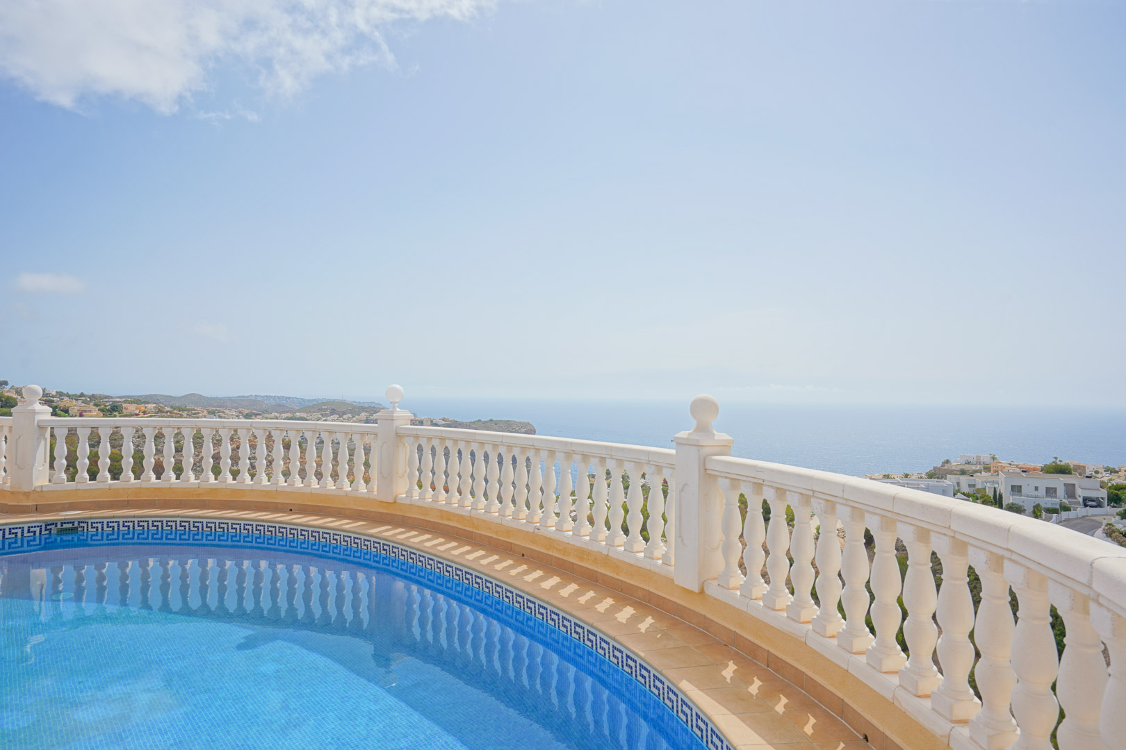 Curved pool with blue mosaic tiles and Greek key border, framed by a white balustrade overlooking the Mediterranean Sea and coastal cliffs.