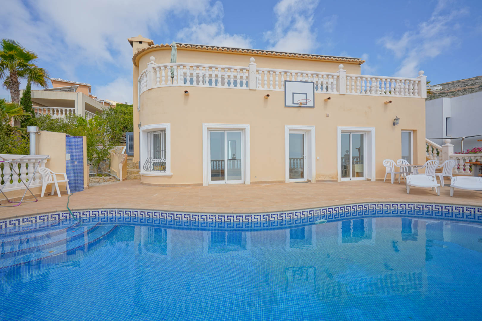 Curved villa facade featuring a blue mosaic pool, terracotta sun terrace with lounge seating, white balustrades, and an integrated basketball hoop.