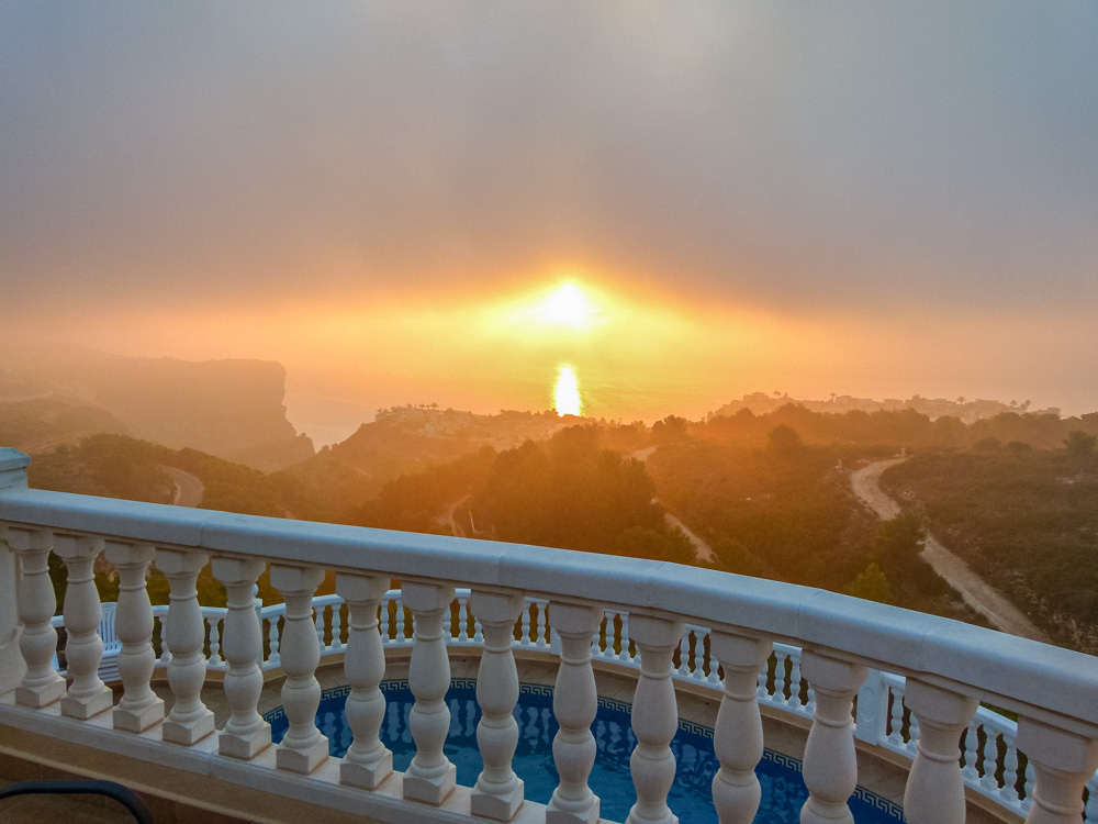 Elevated view from the terrace with white stone balustrades, overlooking the swimming pool and a hazy sunset over the coastal cliffs.