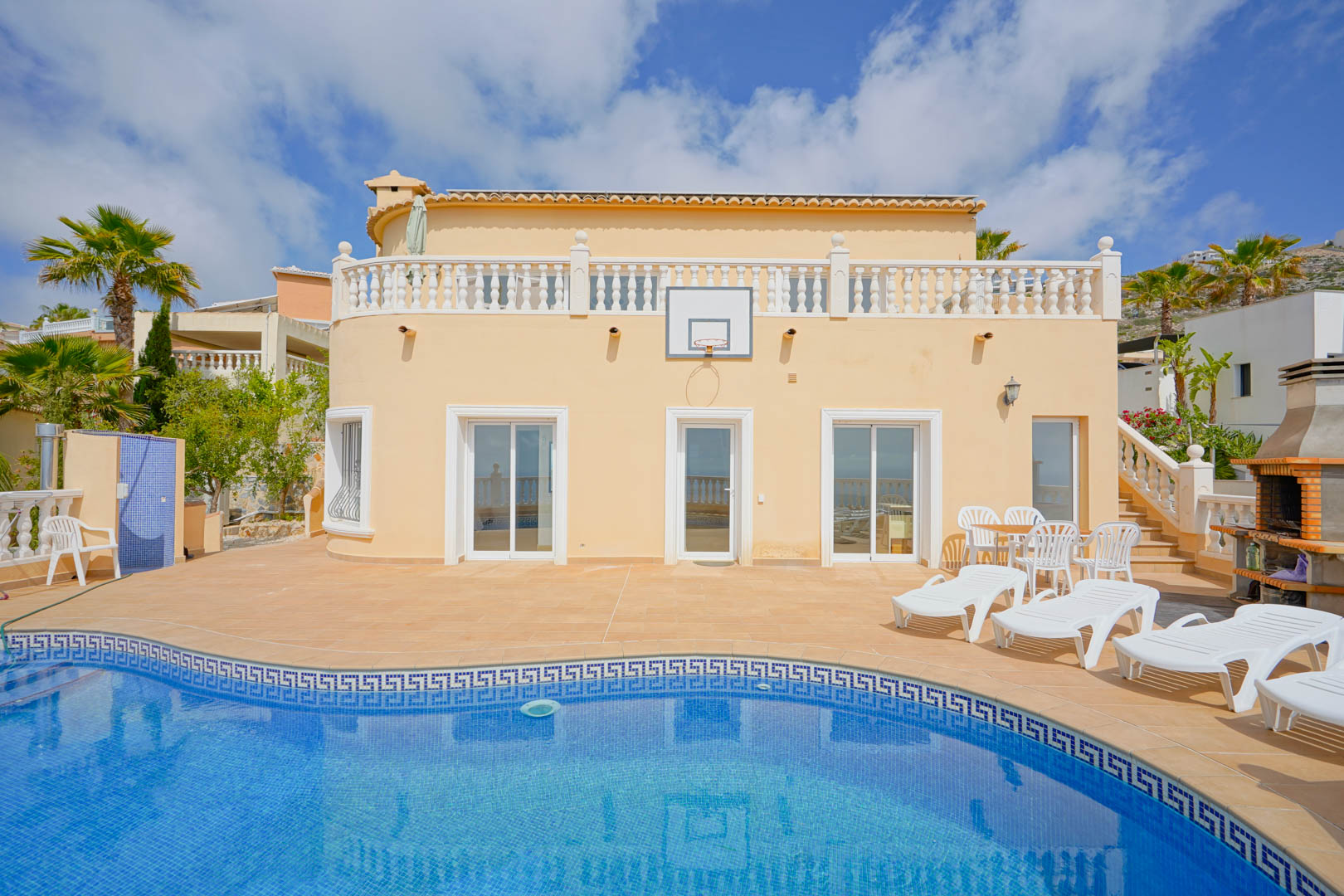 Exterior view featuring a curved swimming pool with mosaic detailing, a spacious sun terrace with loungers, a built-in barbecue, and a basketball hoop.