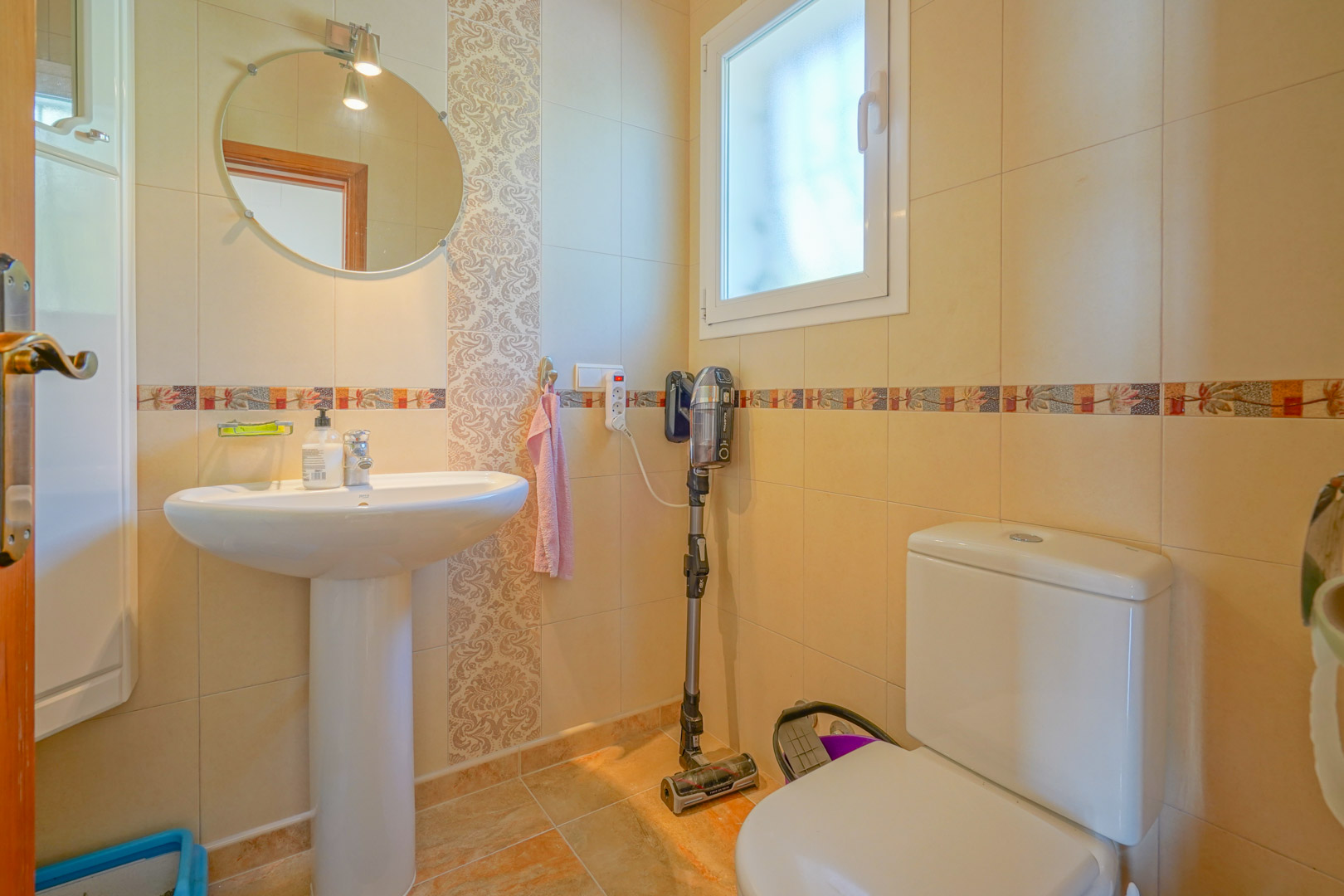Functional bathroom featuring a pedestal sink, circular mirror, and cream-toned tiled walls with decorative borders and a frosted window.