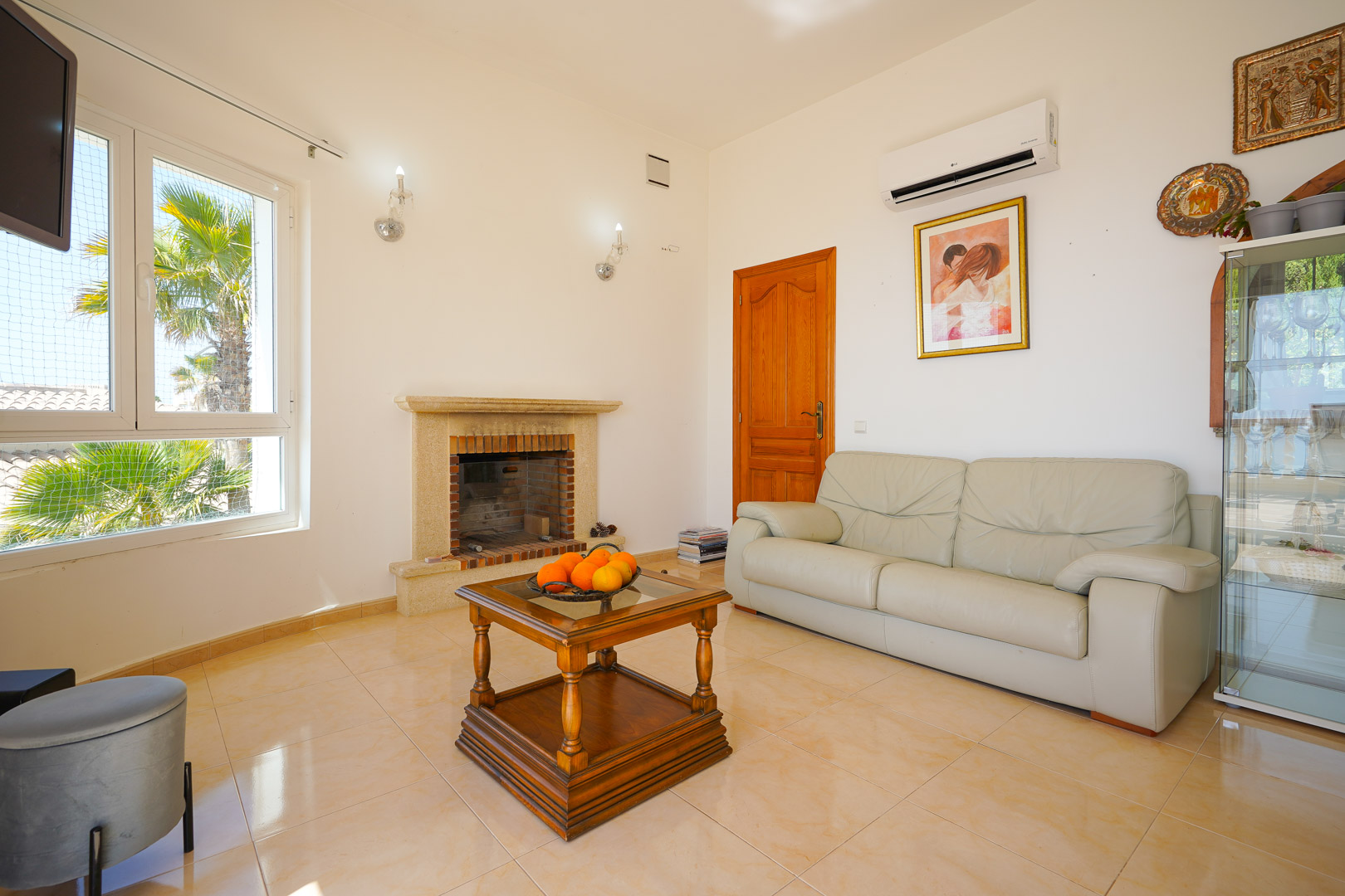 Living area featuring a stone fireplace, beige leather sofa, and wooden coffee table. The space includes an air conditioning unit and views of palm trees through the window.