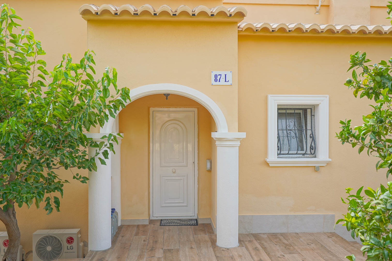 Main entrance featuring a white arched porch with columns, ochre facade, and wood-effect floor tiles, framed by Mediterranean trees and a classic barred window.