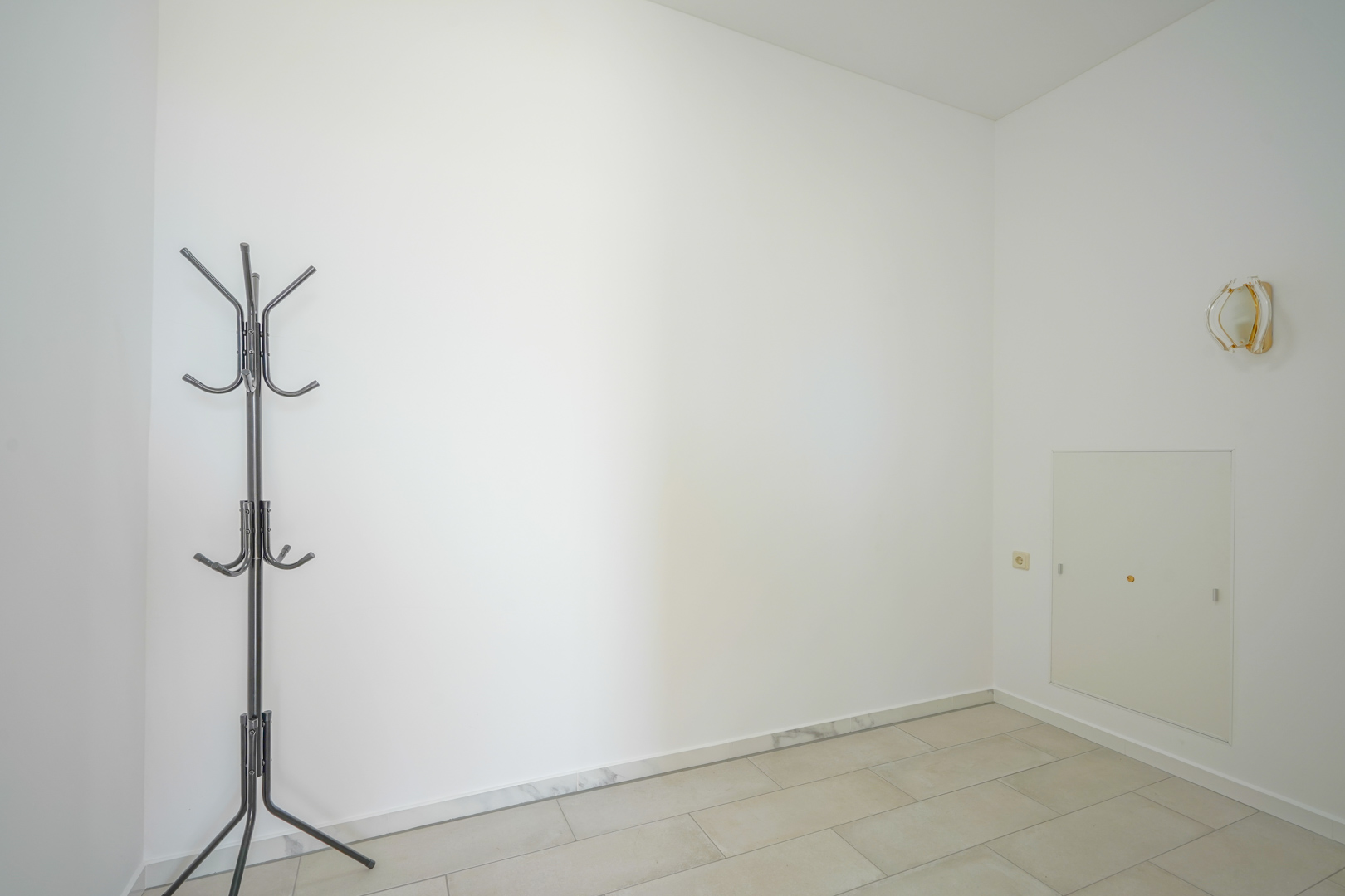 Minimalist entrance area featuring white walls, light ceramic flooring with marble skirting, a black metal coat rack, and a decorative wall sconce.