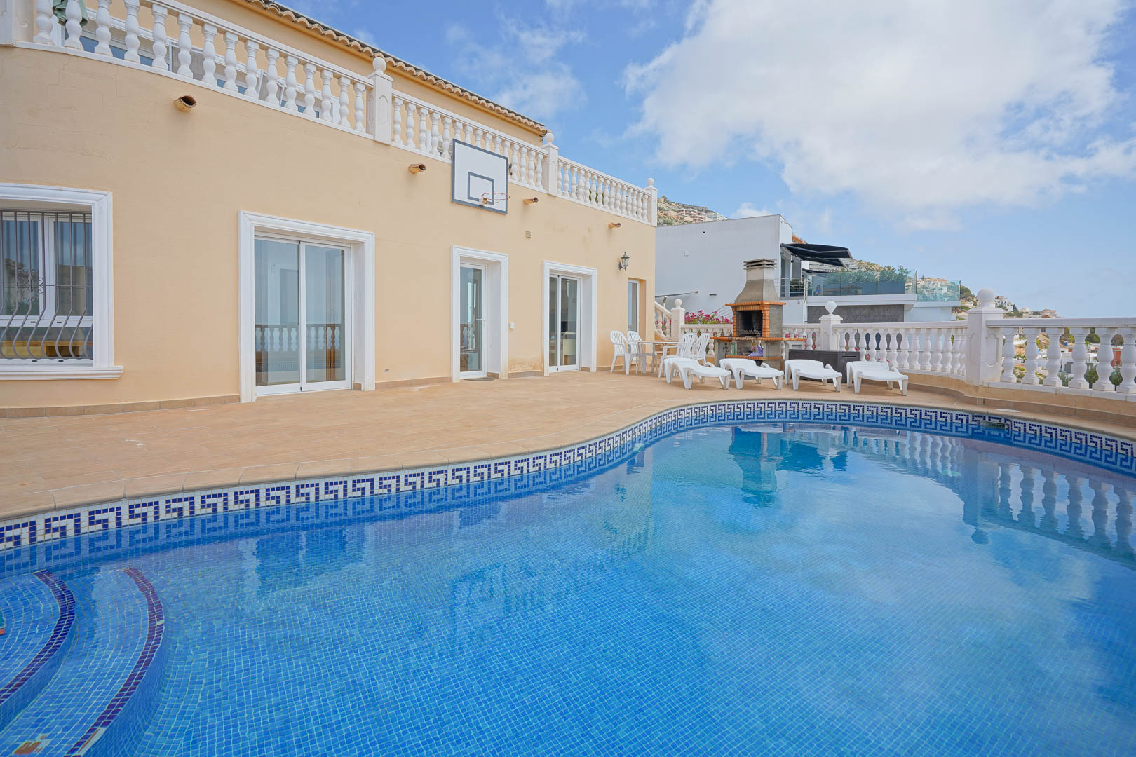 Outdoor terrace featuring a curved pool with blue mosaic tiling and a Greek key border. Includes a built-in stone barbecue, sun loungers, and a basketball hoop.