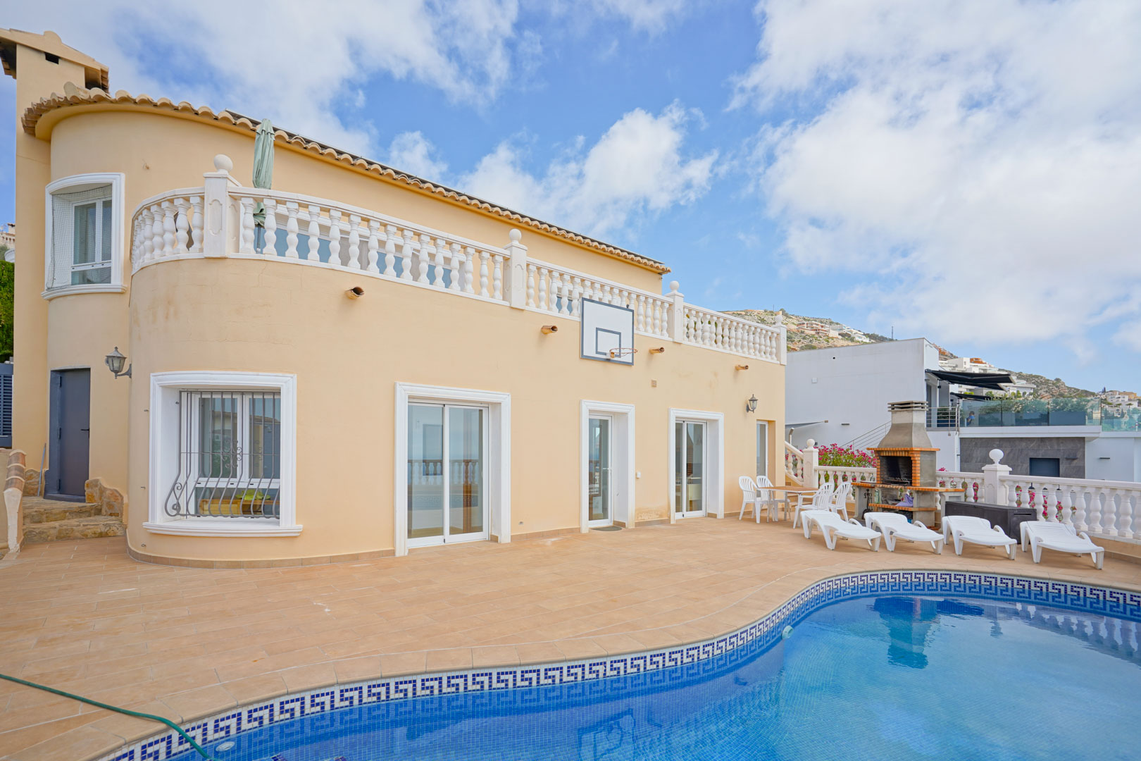 Outdoor terrace featuring a swimming pool with mosaic tiling, a built-in brick barbecue, and sun loungers. The curved villa facade includes white-framed windows and a classic balustrade.