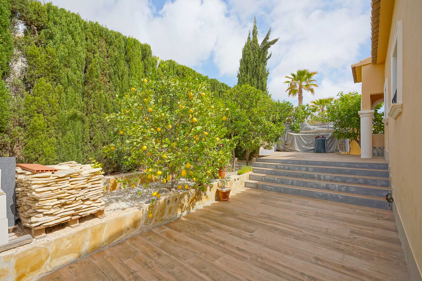 Outdoor terrace featuring wood-effect ceramic tiling, lemon trees, and high cypress hedges, with stone steps connecting the various garden levels.