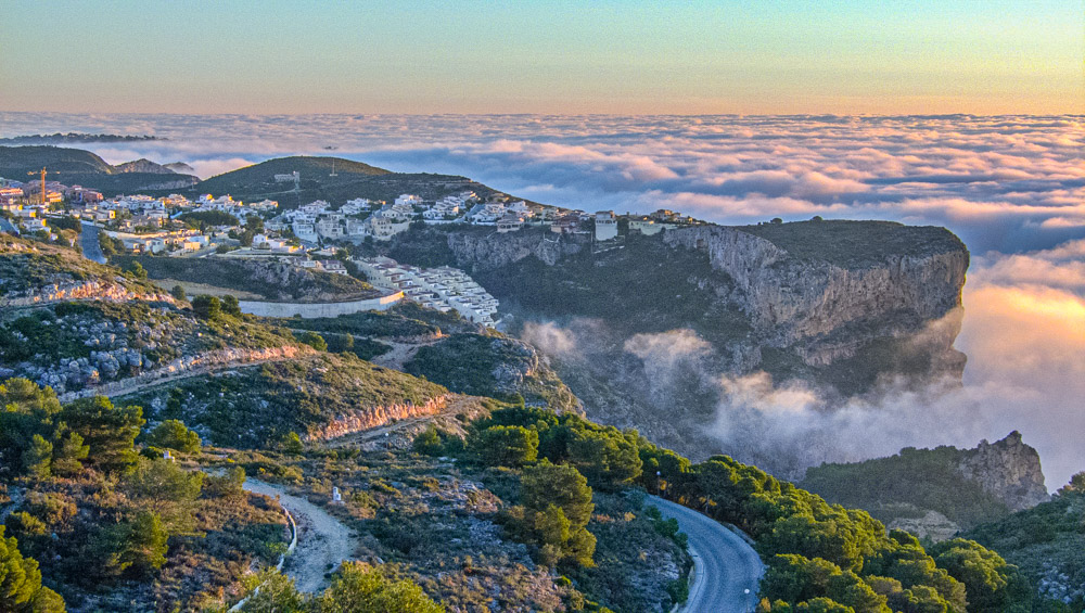 Panoramic view of the Cumbre del Sol cliffs above a sea of clouds at sunset, showcasing the rugged coastline and hillside residential developments.