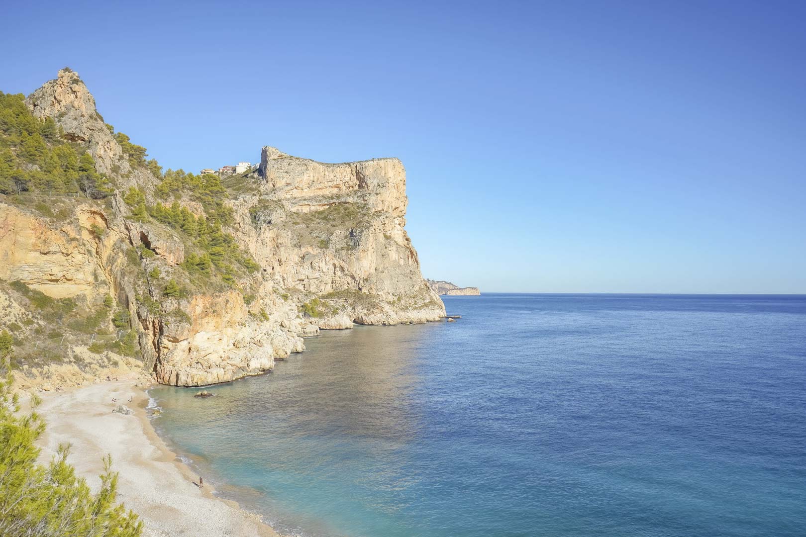 Panoramic view of the Cumbre del Sol cliffs and Cala Moraig beach, featuring the deep blue Mediterranean Sea and rugged limestone formations under a clear sky.