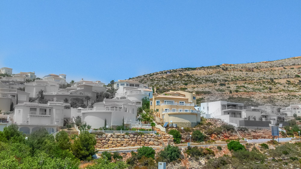 Panoramic view of tiered white villas on the Cumbre del Sol hillside, featuring natural stone retaining walls and curved terraces under a clear sky.