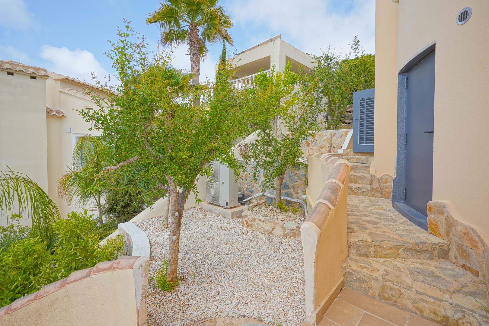 Side terrace featuring fruit trees and white gravel, with natural stone pathways and stairs leading to the upper levels of the villa.