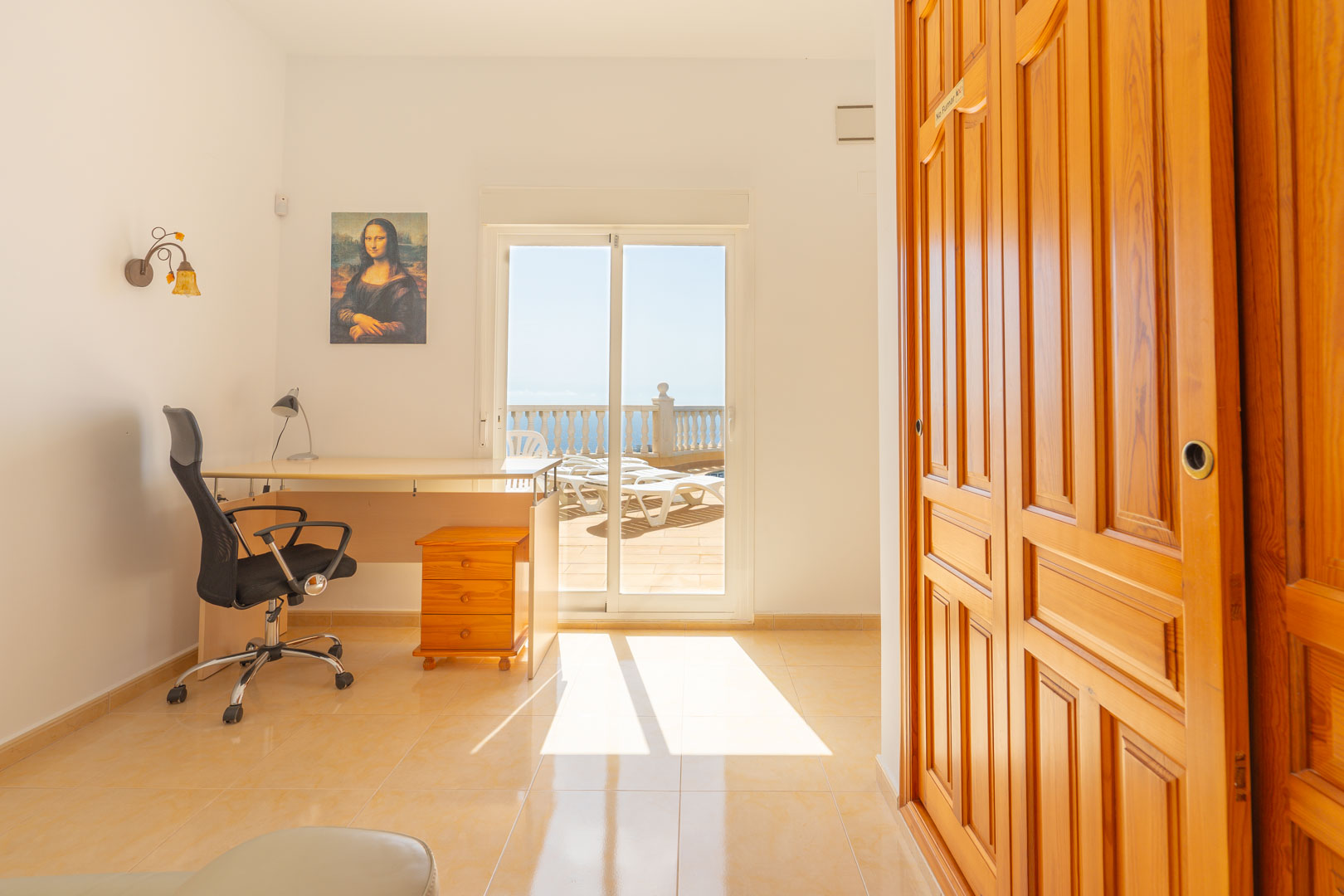 Versatile room featuring a workspace, built-in wooden wardrobes, and direct access to the sun-drenched terrace with coastal views through sliding glass doors.