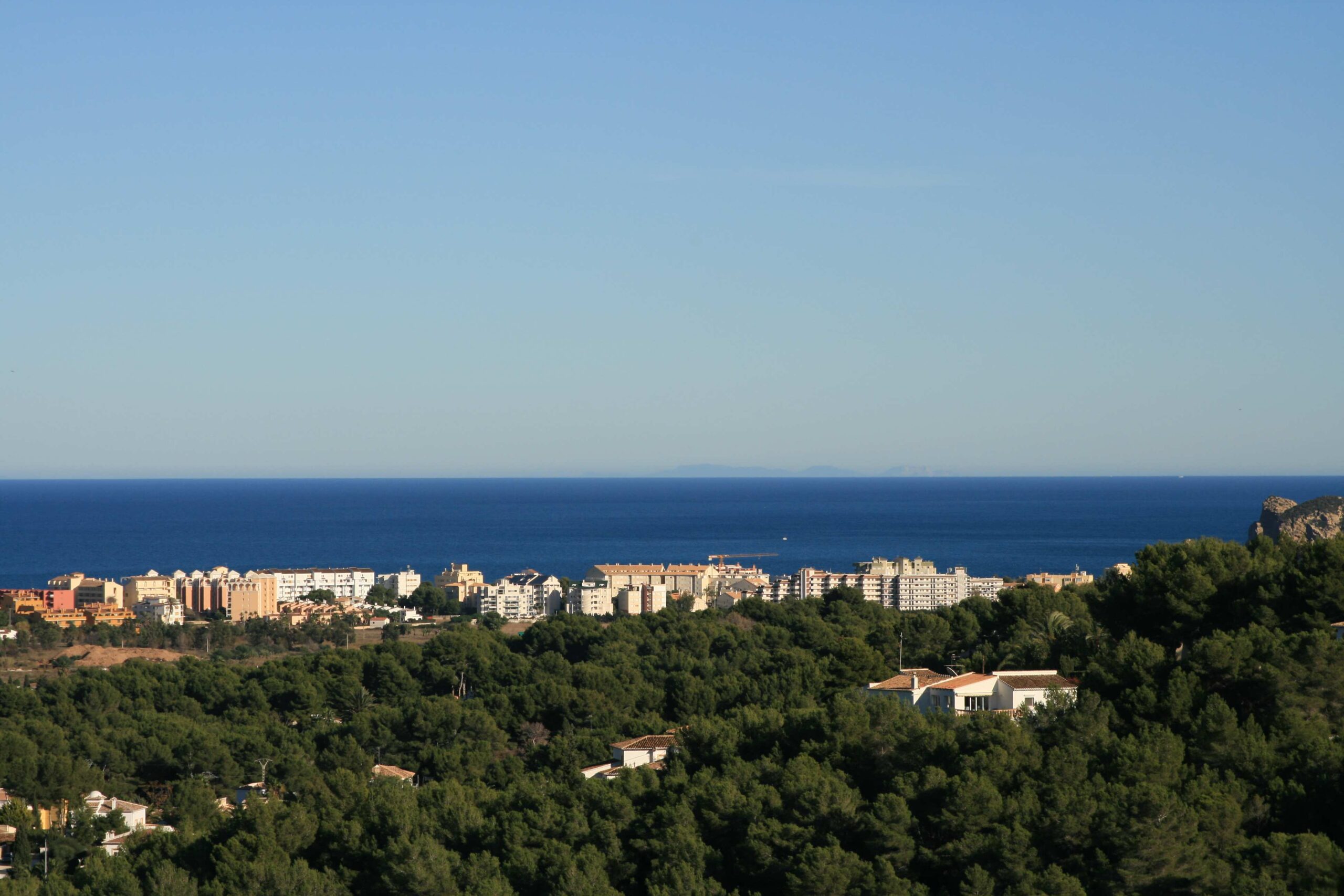 Elevated panoramic view featuring a dense pine forest in the foreground, overlooking a coastal town and the Mediterranean Sea with a clear horizon.