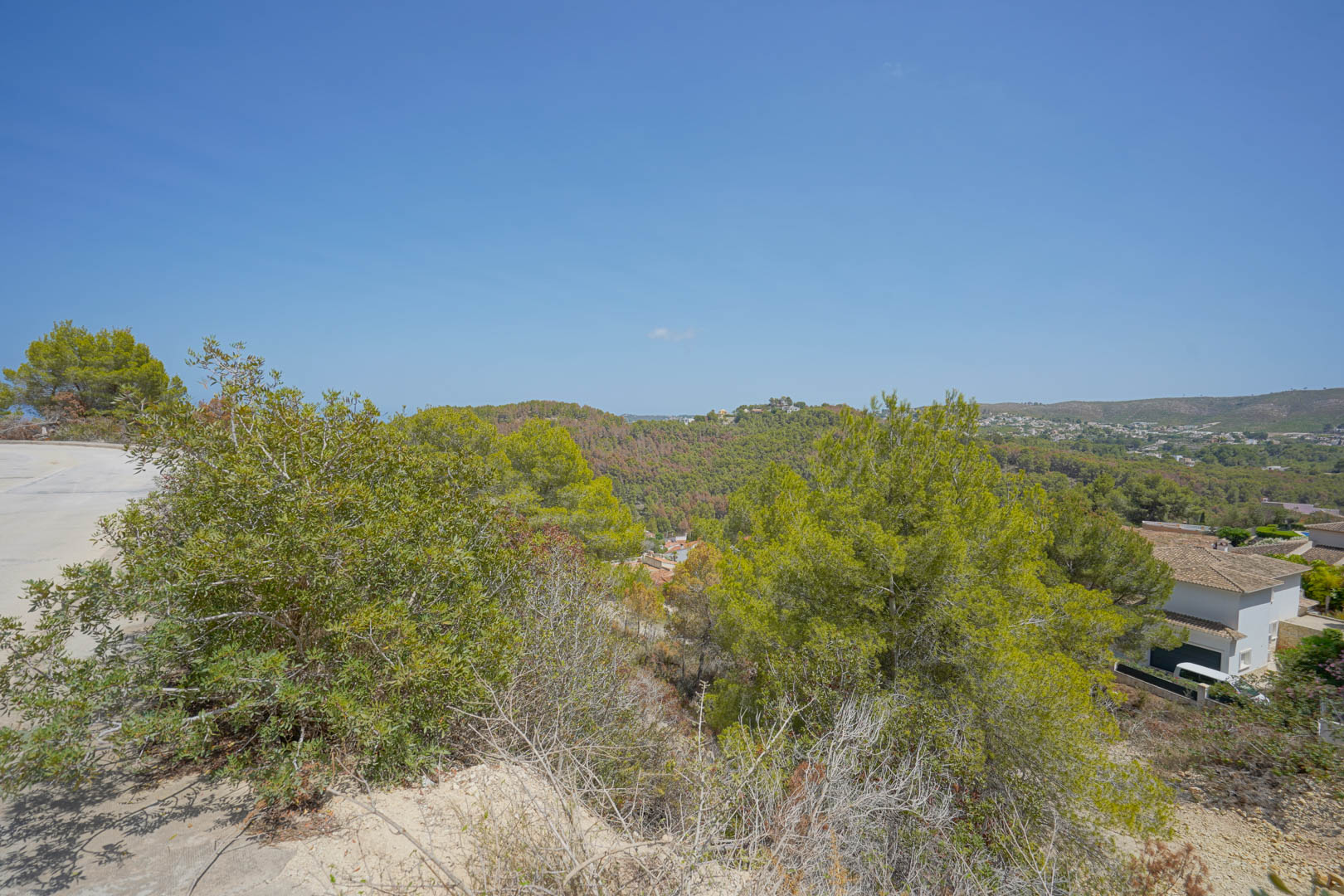 Elevated view from the plot featuring paved road access and panoramic vistas of the surrounding green hills and Mediterranean vegetation.