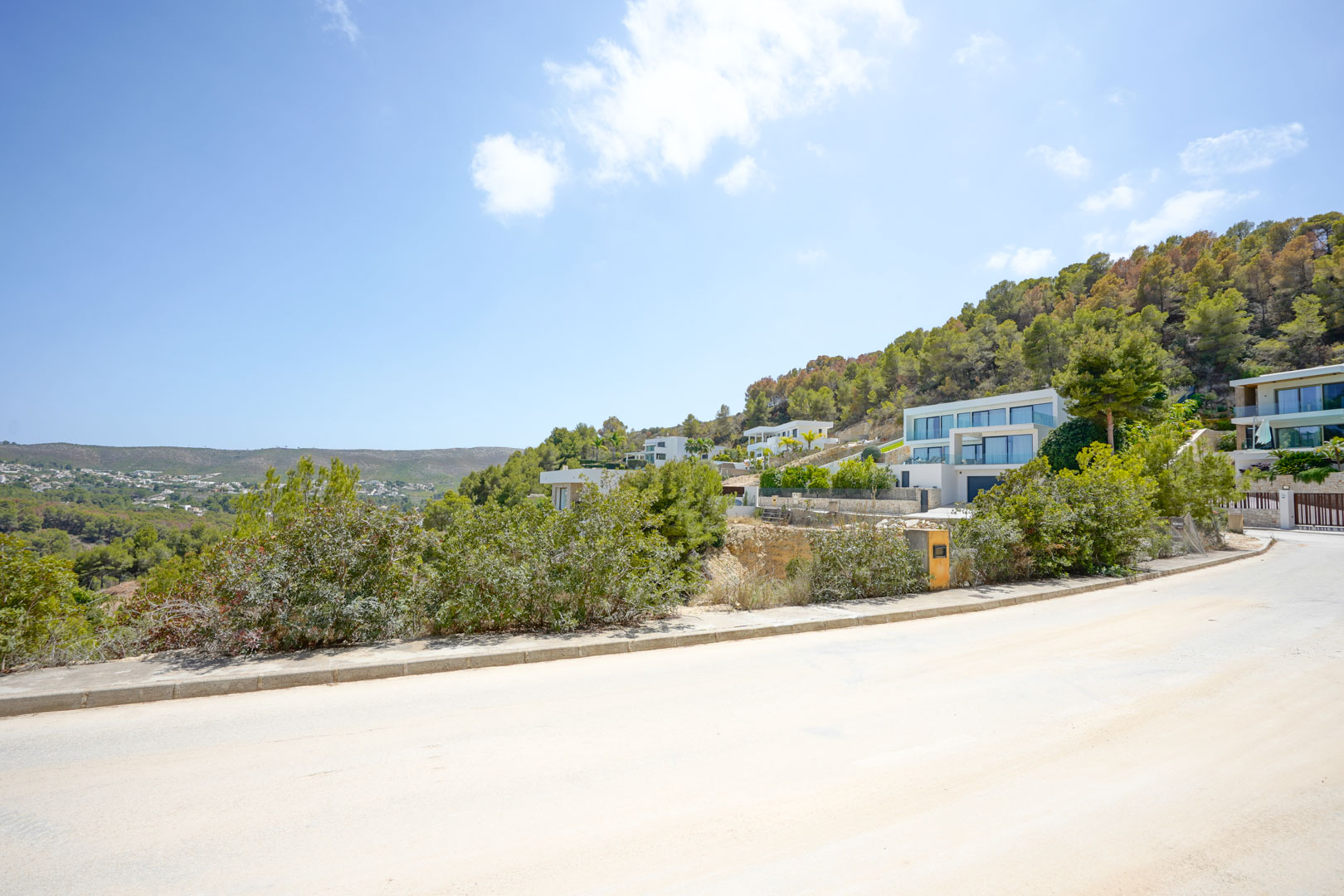 Street-level view of the paved road and sidewalk within the development, featuring modern villas nestled into the steep, pine-covered hillside.