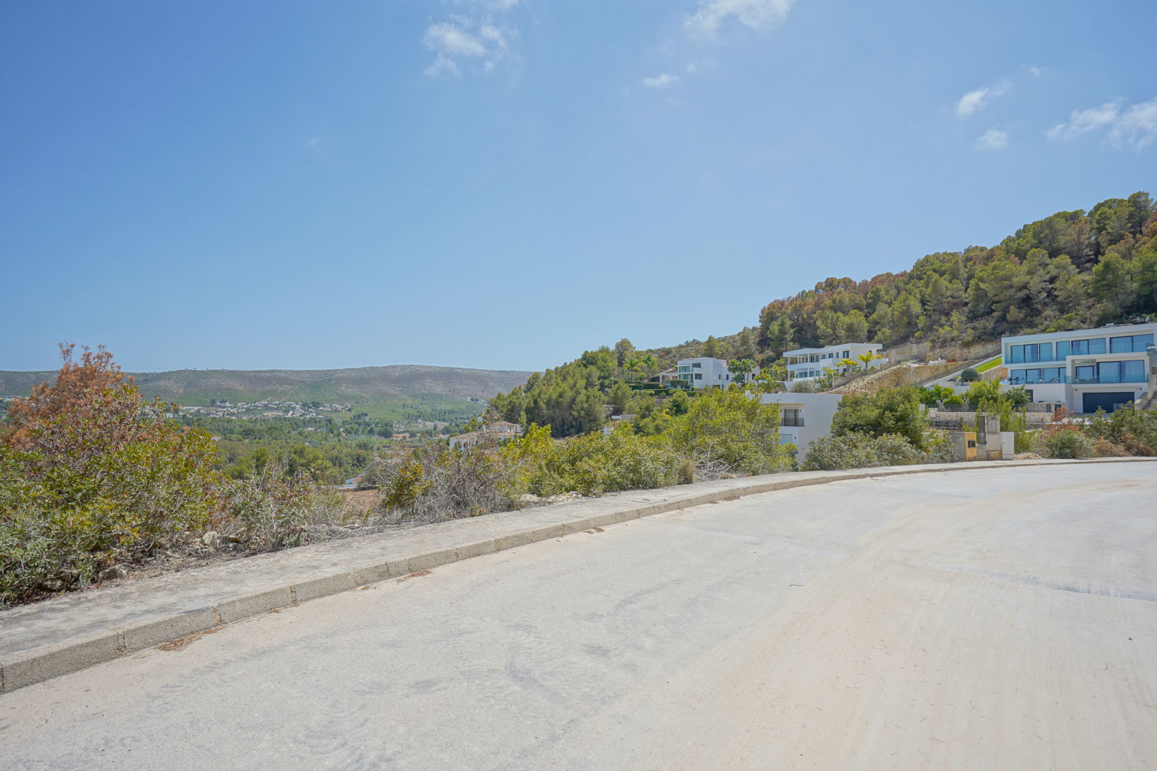 View from the paved access road showing the sloped topography and modern villas integrated into the forested hillside under a clear sky.