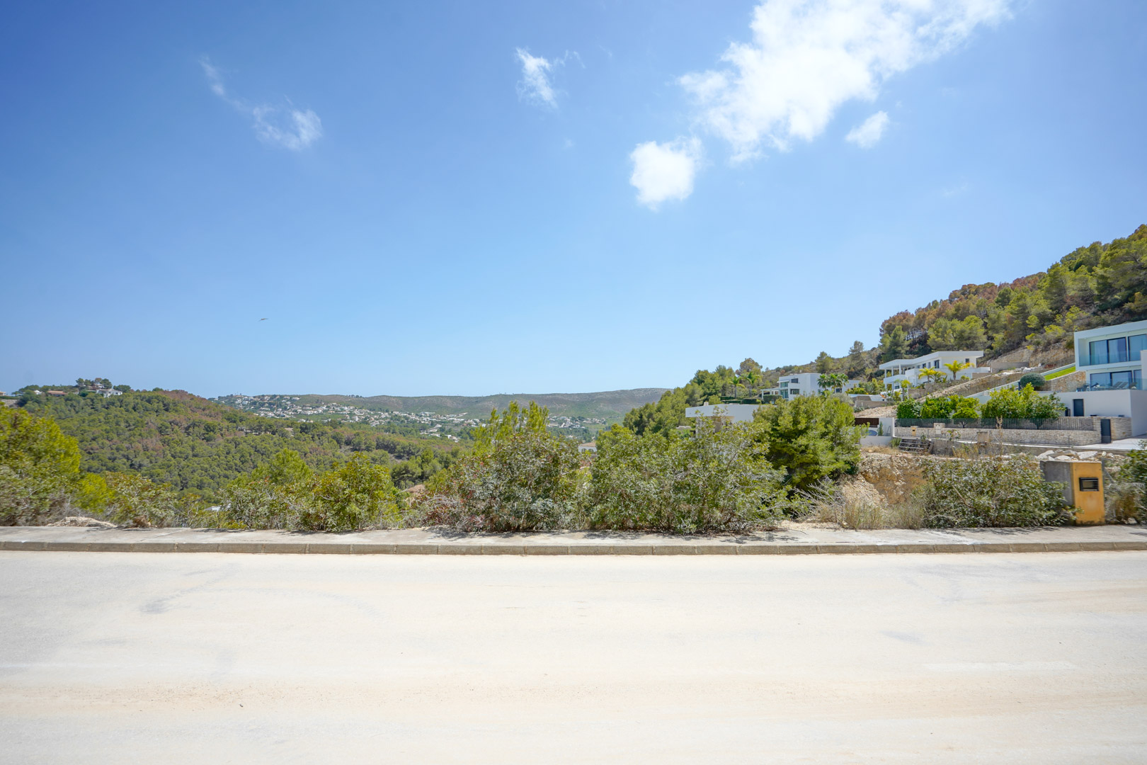 View of the paved access road with concrete curbing in a modern development, highlighting the elevated position and surrounding green hillsides.