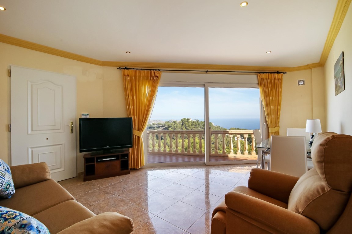 Bright living area featuring tiled flooring, crown molding, and large sliding doors that open to panoramic views of the Mediterranean Sea and lush hills.