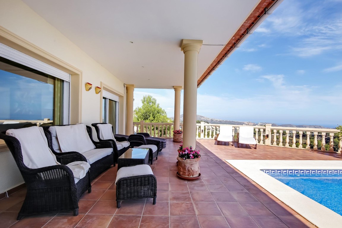 Covered terrace featuring classic columns, wicker seating, and terracotta tiling adjacent to the pool, overlooking the Mediterranean Sea and hills.