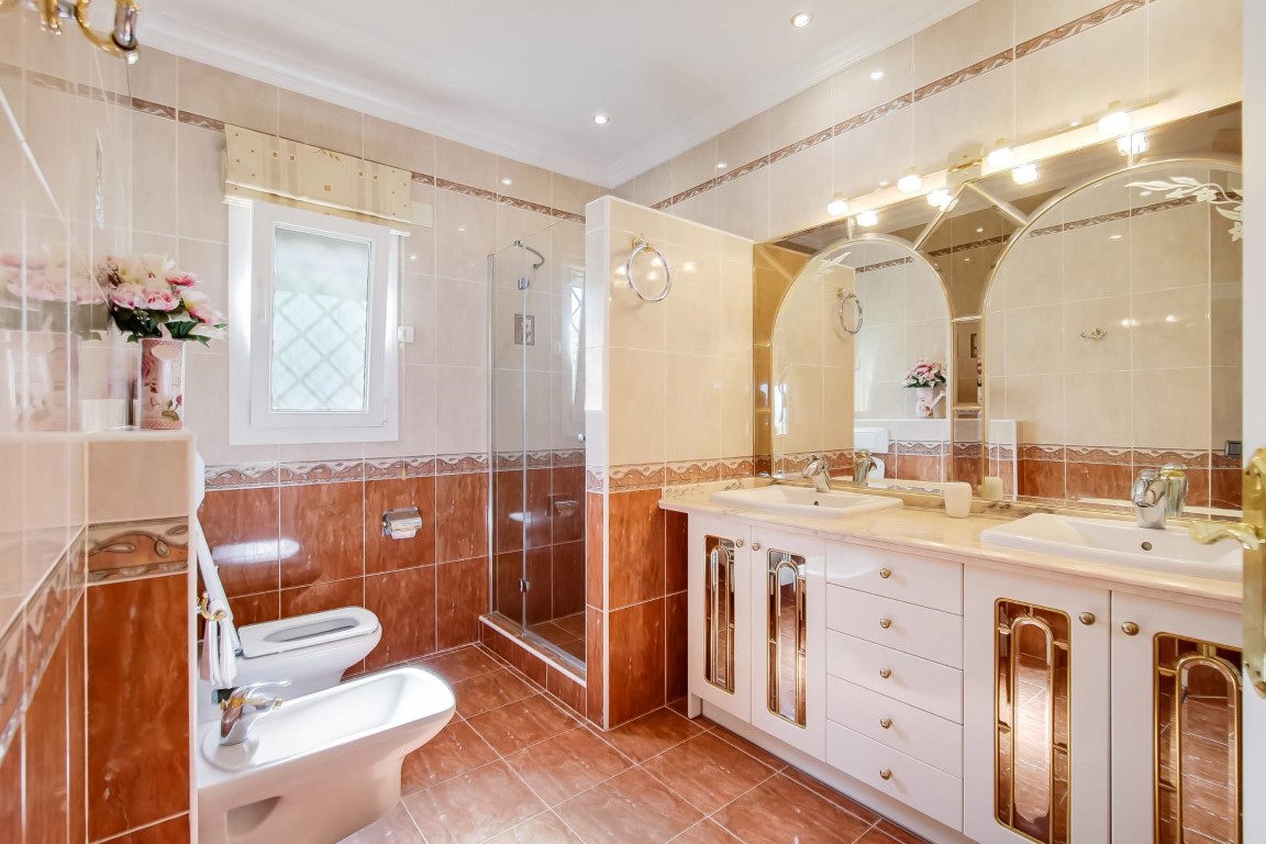 Full bathroom featuring a double vanity with gold accents, arched mirrors with integrated lighting, and a walk-in glass shower.