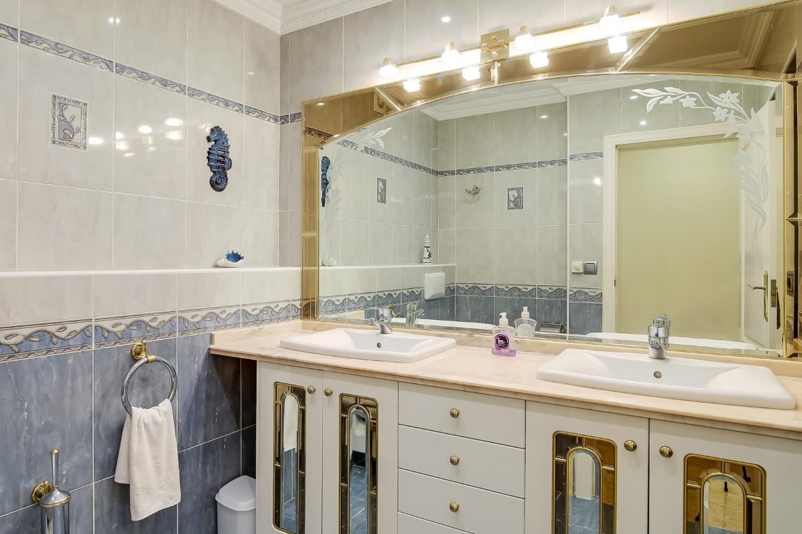 Main bathroom featuring a double vanity with marble countertop, gold-trimmed cabinetry, a large illuminated mirror, and blue decorative tiling.