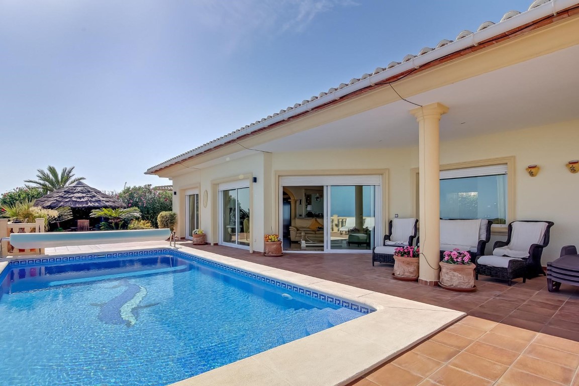 Main terrace featuring a mosaic-inlaid pool, covered porch with columns, and outdoor seating. A thatched-roof gazebo is visible amidst the Mediterranean landscaping.