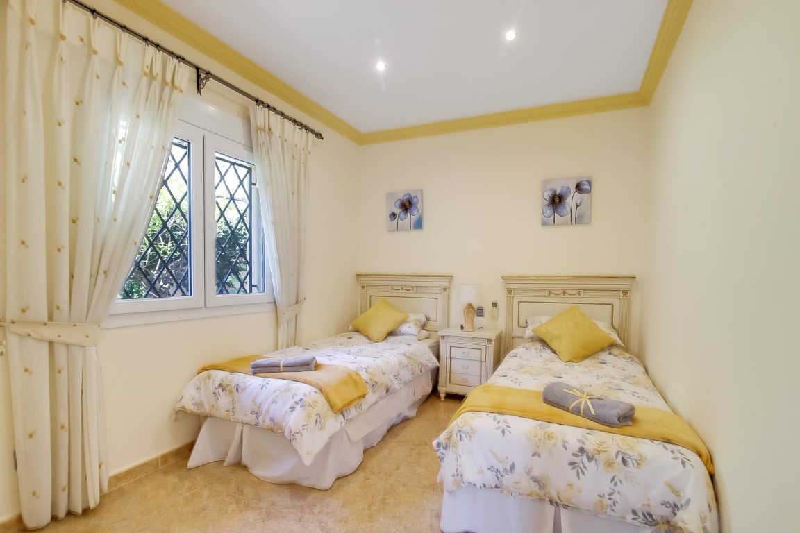 Twin bedroom featuring white wooden headboards, recessed ceiling spotlights, and a large window with security grilles and light curtains.