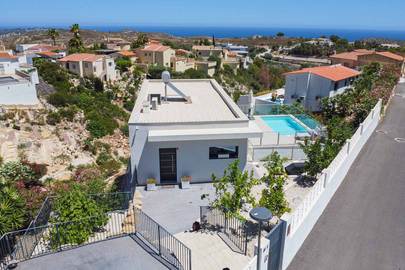 Aerial view showing the modern villa's flat roof with solar heating, the private pool area with glass railings, and the surrounding Mediterranean landscape.