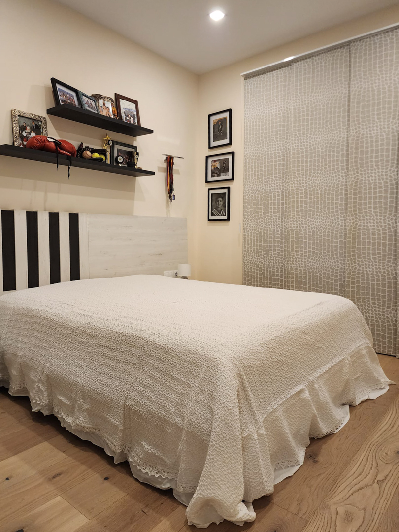 Bedroom featuring light oak flooring, a custom headboard, and floating shelves. The space includes textured panel curtains and recessed ceiling lighting.