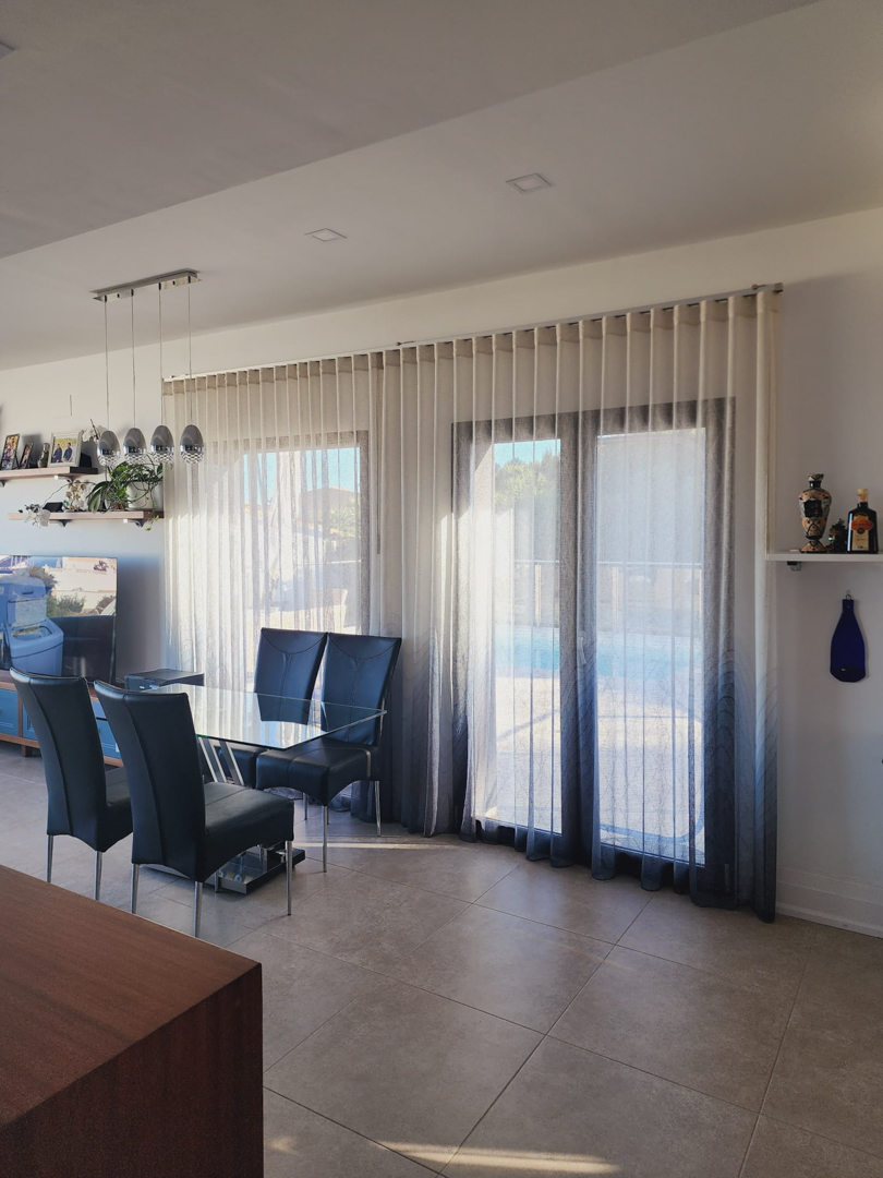 Bright dining area featuring a glass table and high-back chairs, positioned next to large windows with gradient curtains offering direct access to the pool terrace.