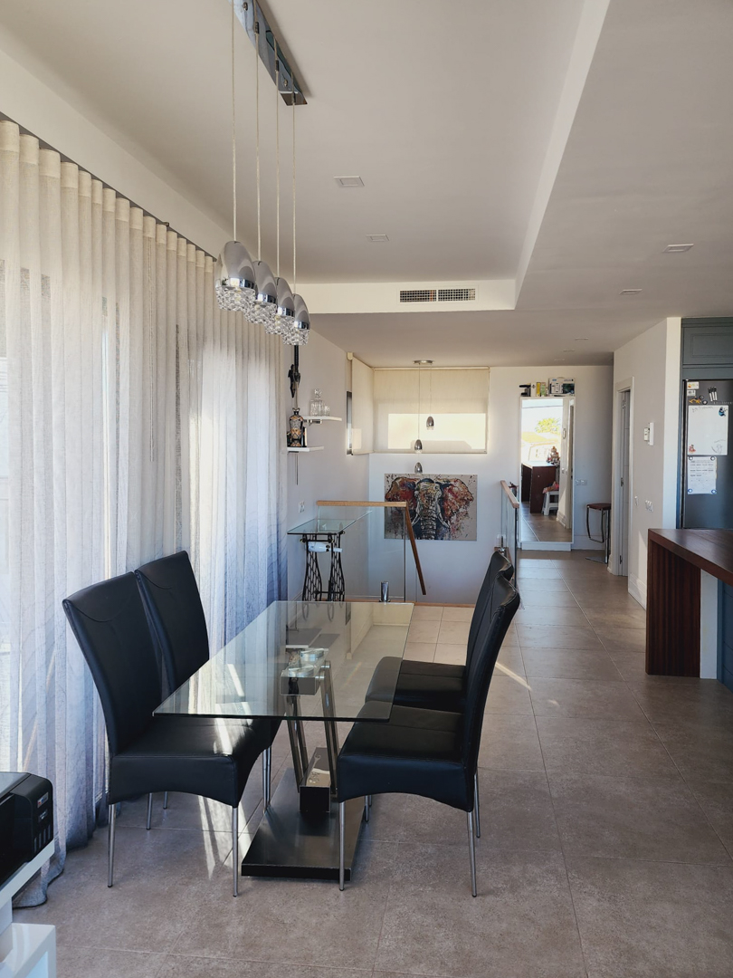 Bright dining area featuring a glass table, black leather chairs, and modern pendant lighting, situated next to large windows with sheer curtains and a staircase.