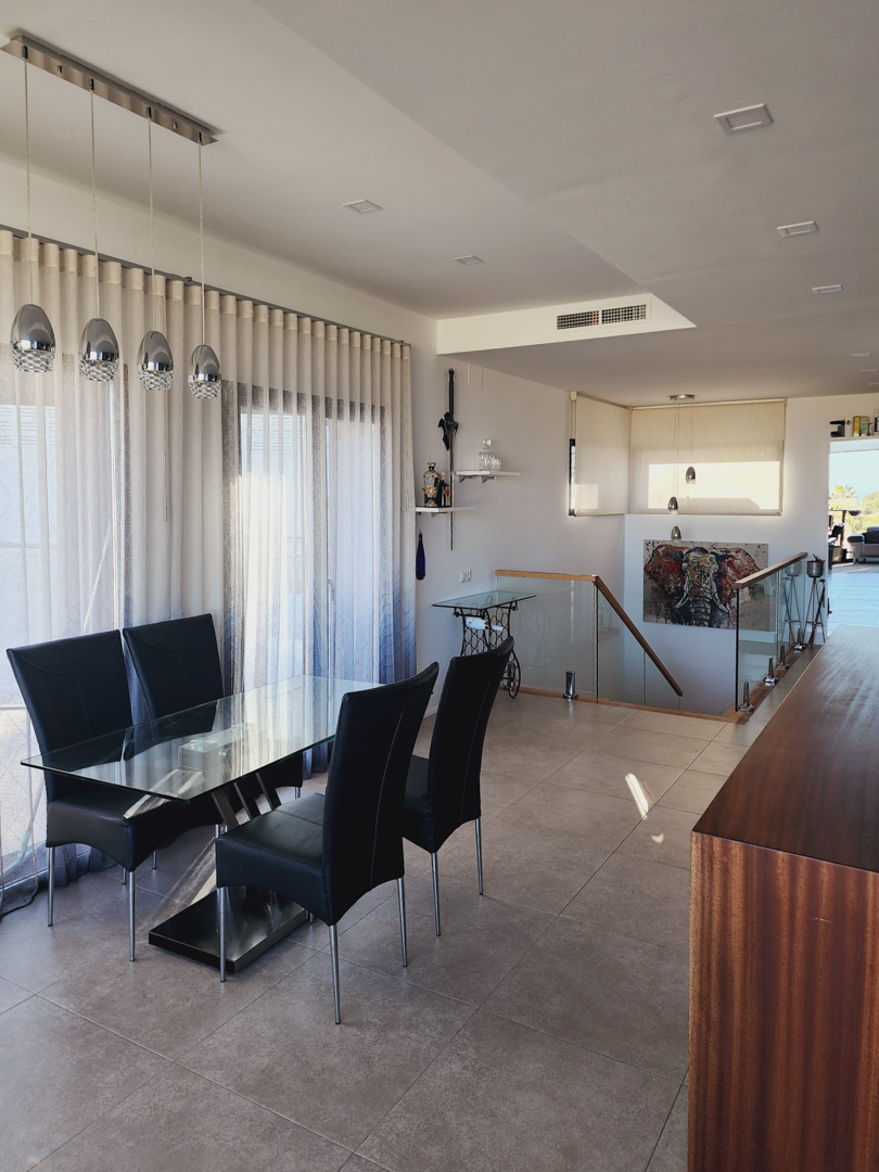 Dining area featuring a glass-top table with black chairs, situated next to a staircase with a glass balustrade. Modern pendant lighting and recessed ceiling fixtures illuminate the space.