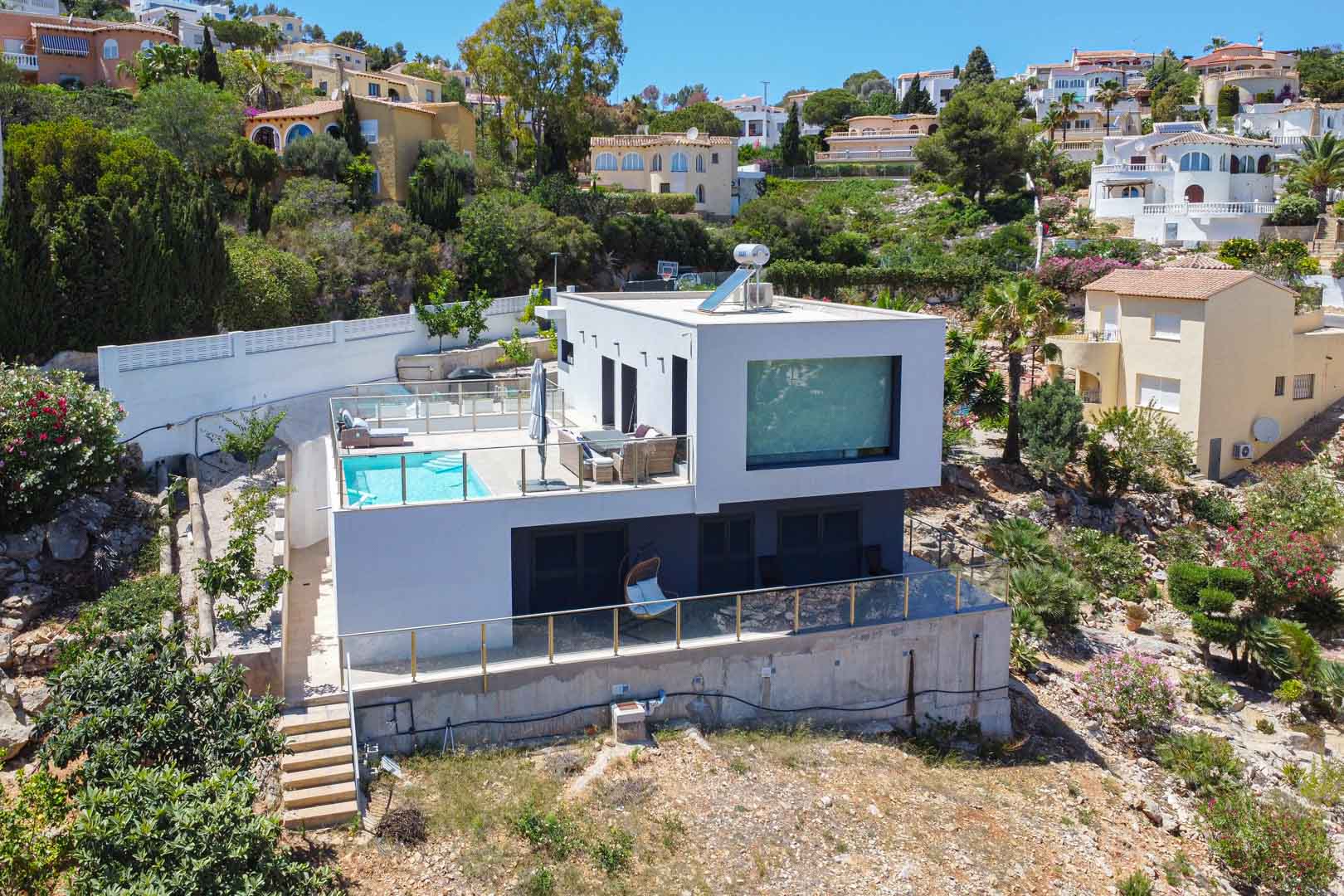 Exterior view of the modern cubic villa featuring an upper terrace with a swimming pool, glass railings, and a solar water heater on the flat roof.