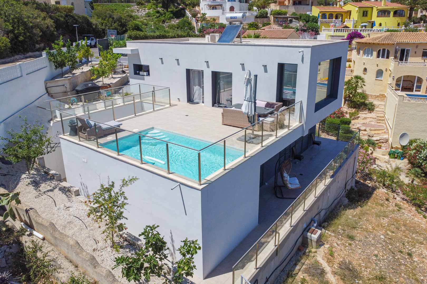 Exterior view of the modern villa featuring an upper terrace pool with glass balustrades, rooftop solar panels, and a secondary covered terrace below.