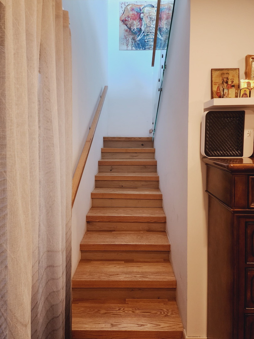 Interior wooden staircase featuring a matching handrail and glass balustrade, illuminated by natural light from above and accented by wall art.