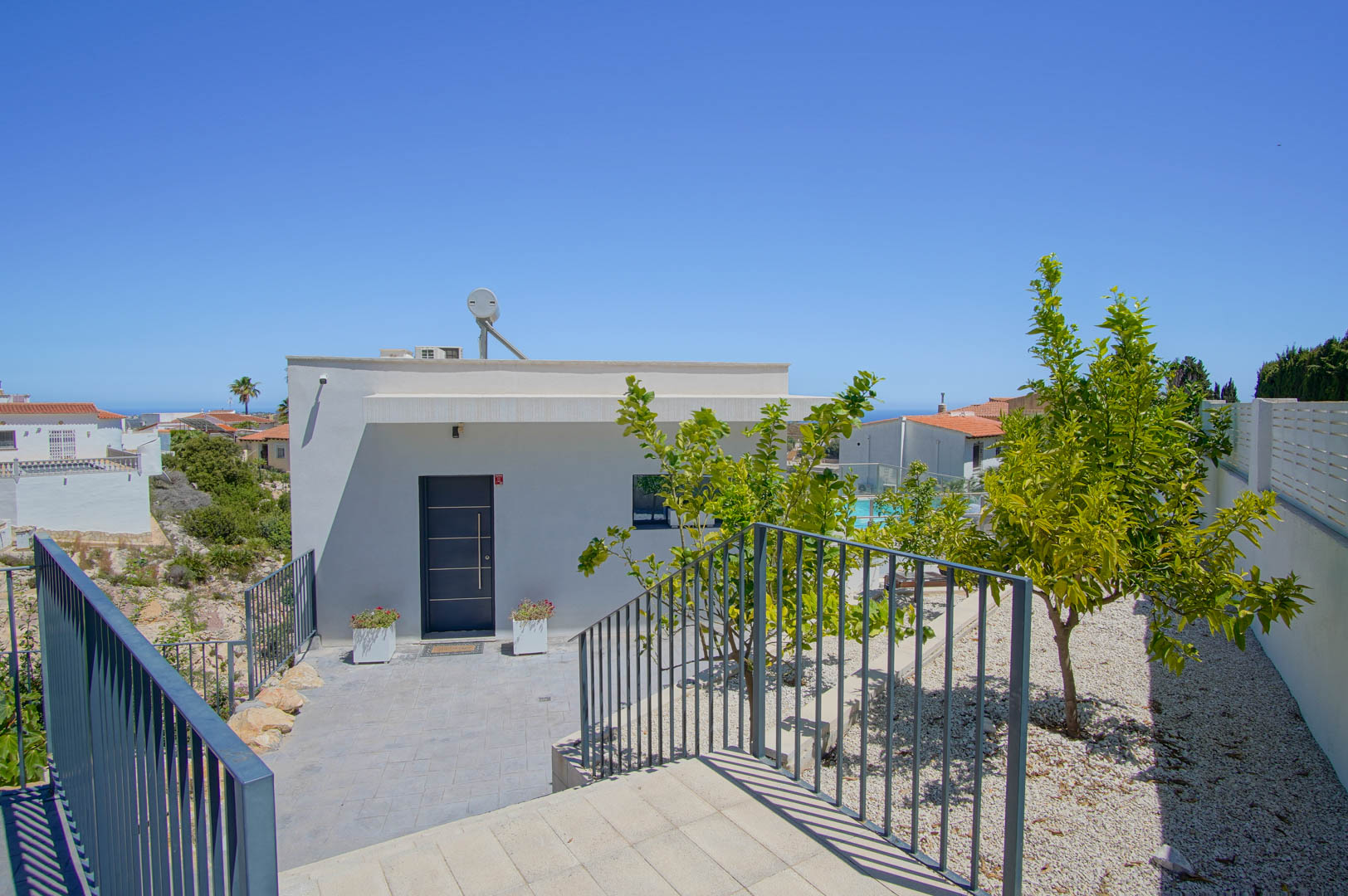 Main entrance featuring a modern white facade, dark grey security door, solar water heater, and a gravel garden with young citrus trees.