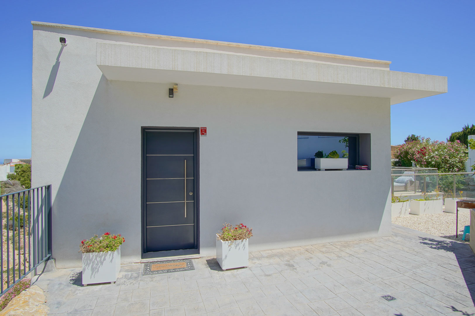 Minimalist white facade featuring a dark grey modern entrance door with a vertical steel handle and decorative planters on the paved stone entryway.