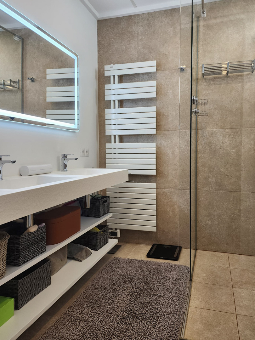 Modern bathroom featuring a double integrated vanity, open shelving, and an LED-backlit mirror. It includes a white towel radiator and a walk-in shower with earth-toned tiling.