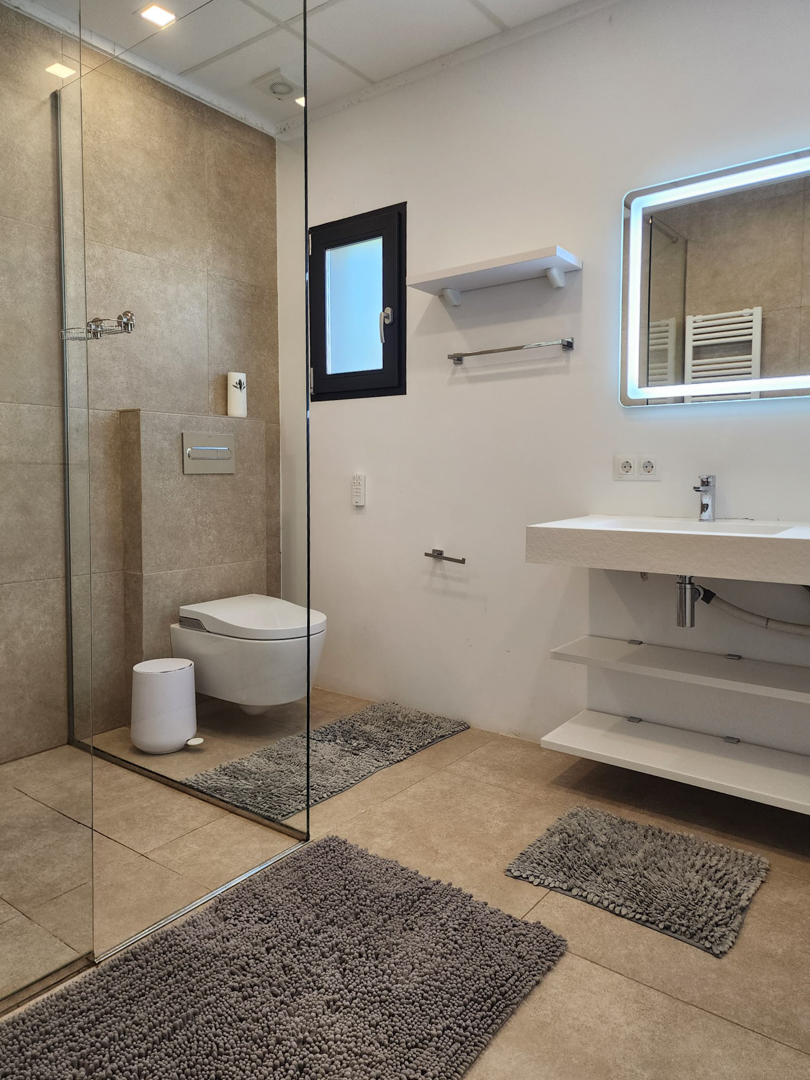 Modern bathroom featuring a walk-in glass shower, wall-mounted toilet, and a floating vanity with open shelving and a backlit LED mirror against beige tiling.