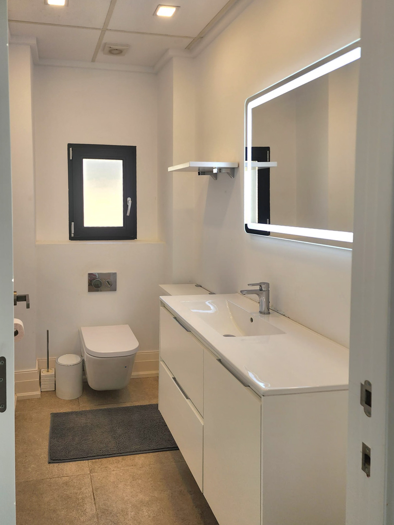 Modern bathroom featuring a white vanity unit, LED backlit mirror, and wall-mounted toilet. It includes recessed ceiling lighting and neutral stone-effect floor tiles.
