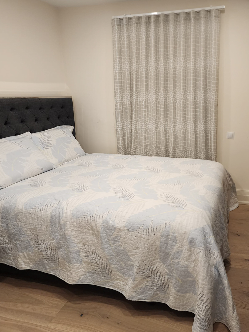 Modern bedroom featuring a double bed with a dark grey tufted headboard, light wood flooring, and floor-to-ceiling patterned curtains.