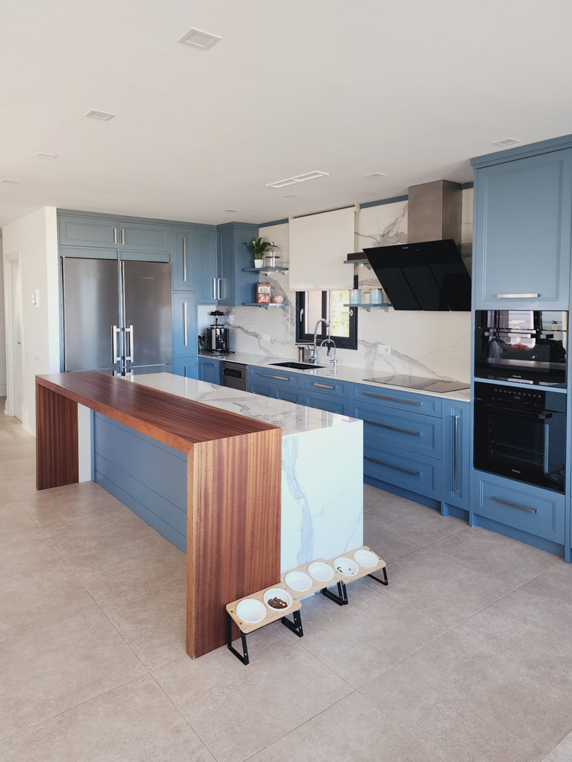 Modern kitchen featuring blue cabinetry, a marble waterfall island with an integrated wood breakfast bar, and high-end Miele appliances against a matching stone backsplash.