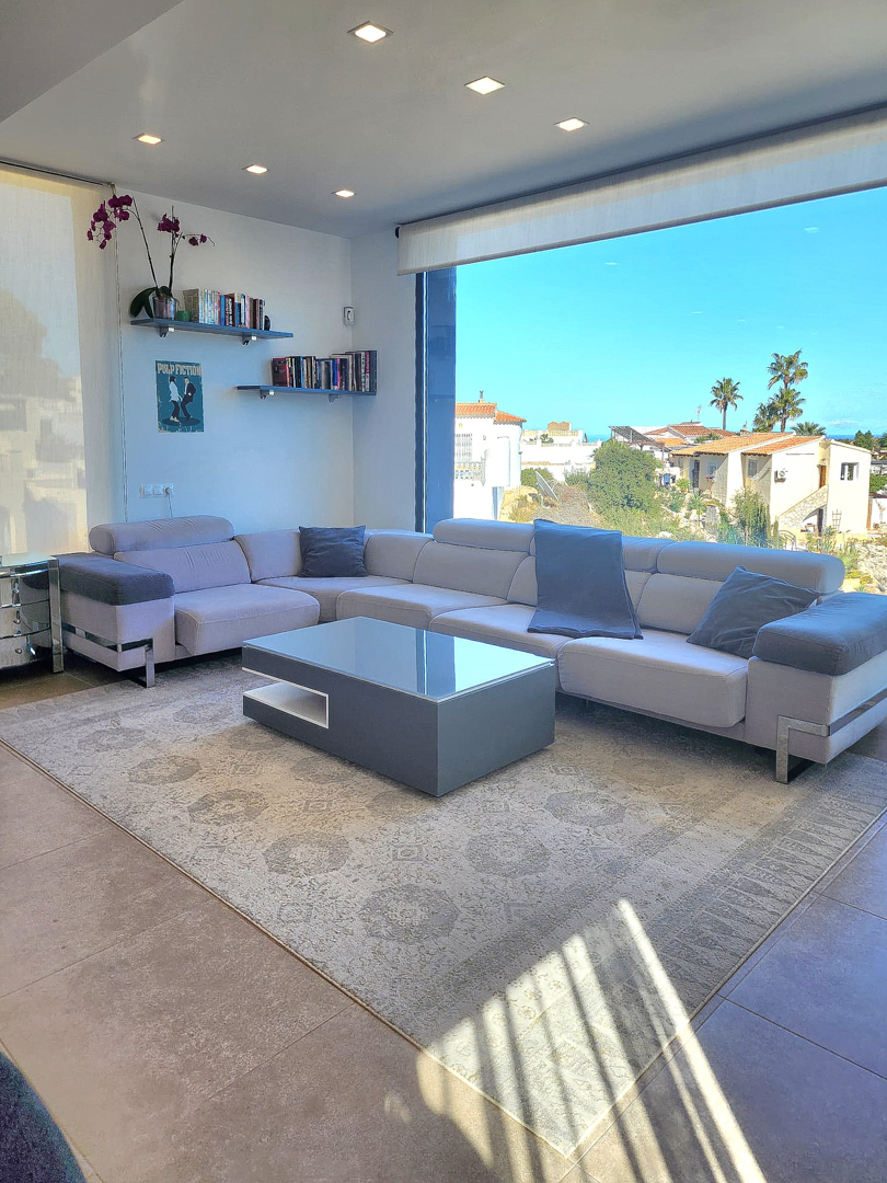 Modern living room with an L-shaped sofa, minimalist coffee table, and large picture window. Features recessed ceiling lights, floating shelves, and porcelain tile flooring.