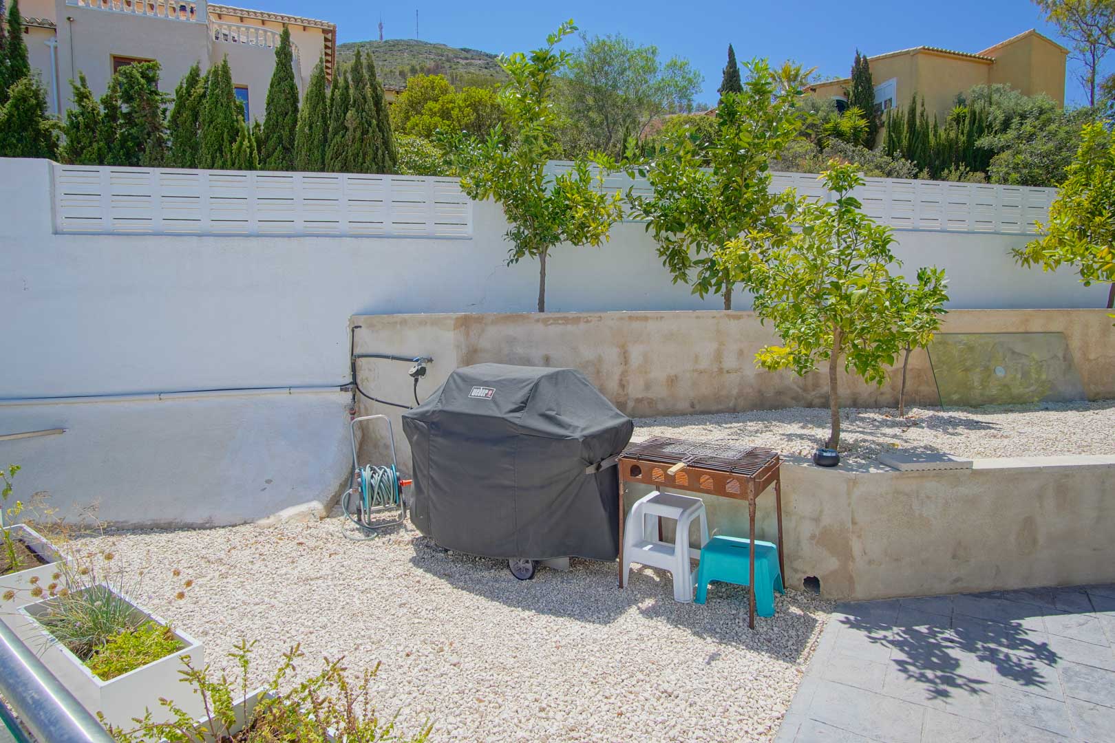 Outdoor garden area with white gravel, young citrus trees, and a barbecue station. The space is enclosed by a white wall under clear Mediterranean skies.