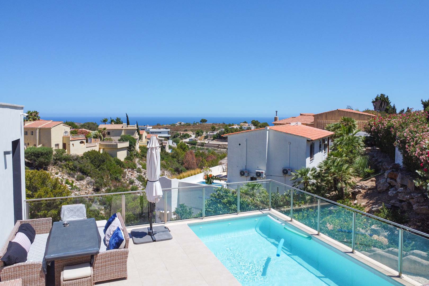 Outdoor terrace featuring a swimming pool with glass balustrades, wicker dining furniture, and panoramic Mediterranean Sea views under a clear sky.