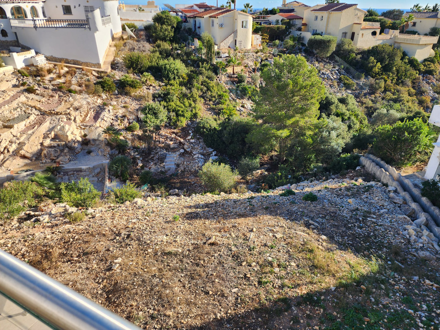Perspective from the balcony showing the sloped plot with gravel terrain, natural rock formations, and native Mediterranean greenery.